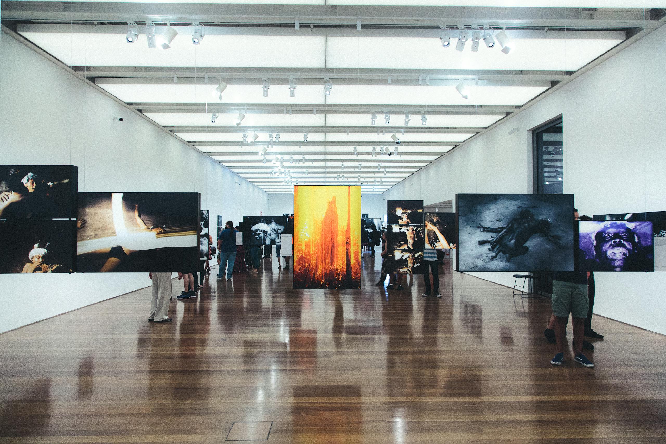 A contemporary art gallery with visitors observing various photographic exhibitions displayed on walls and stands.